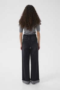 Μαύρο τζιν παντελόνι φαρδιάς γραμμής με ψηλή μέση, γυρισμένα μανσέτες και δύο πίσω τσέπες. Φαίνεται το εφαρμοστό γκρι τοπ του μοντέλου.