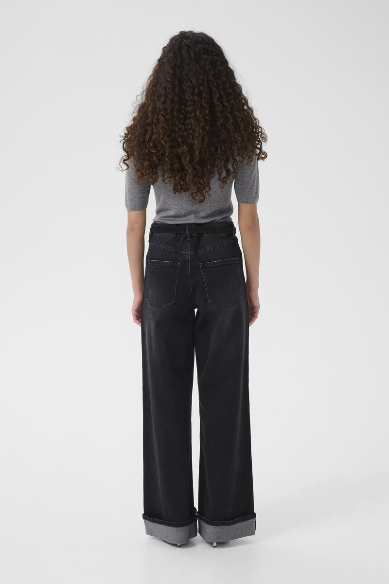 Jeans in denim nero a gamba larga con vita alta, risvolti arrotolati e due tasche posteriori. Si vede la maglietta aderente grigia del modello.