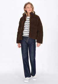 Braune Fleecejacke mit hohem Kragen, getragen über einem weiß-schwarz gestreifen Hemd, kombiniert mit blauen Jeans und weißen Sneakers.