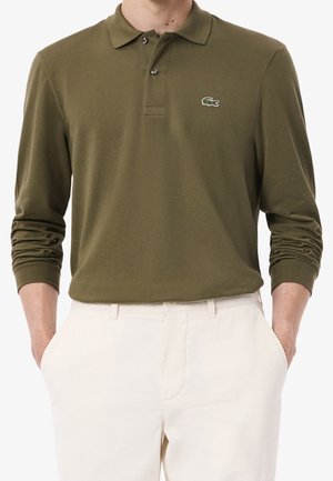 Mann trägt olivgrünes langärmeliges Poloshirt mit Krokodil-Logo und weiße Hose, Hände in den Taschen, steht vor weißem Hintergrund.