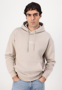 Hanorac bej din material moale, cu buzunar frontal, șnururi și print cu logo. Asortat cu blugi albaștri deschiși, design minimalist.
