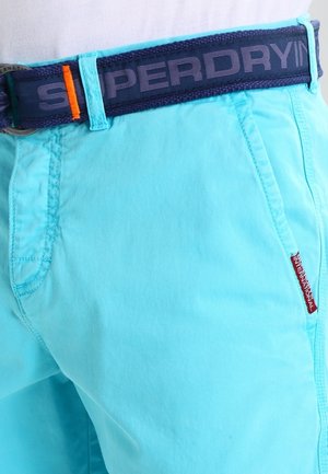 Lyseblå shorts med sidelommer og et navyblåt bælte med teksten "SUPERDRY" og en lille rød mærkat på lommen.