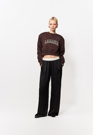 Blondínka stojaca s rukami v vreckách, oblečená v tmavohnedej krátkej mikine s nápisom "LAMANIA" a voľnými čiernymi nohavicami na bielom pozadí.