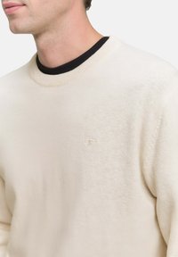 Un pull crème clair avec une texture, un col rond et un petit logo sur la poitrine, superposé sur un T-shirt noir.