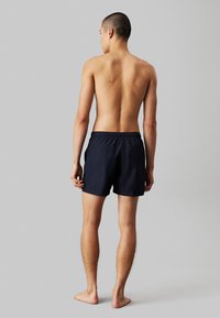 Shorts de bain navy avec taille élastique, intérieur doublé et poches latérales. Tissu lisse et coupe au-dessus du genou. Vue de dos.