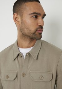 Beige sako s špičatým límcem, obsahující dvě náprsní kapsy na knoflíky a texturovanou látku. Nosené přes bílou košili.