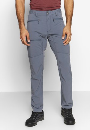 Homme portant un pantalon de plein air gris avec des poches zippées et des chaussures de randonnée noires, debout devant un fond blanc uni.