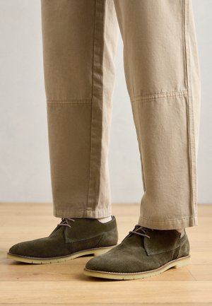 Zapatos de ante verde con una parte superior Texturizada, suela ligera y cordones marrones, combinados con pantalones en un tono beige claro con costuras visibles.