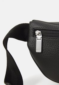 Borsa in pelle nera con superficie texturizzata, hardware della cerniera in argento e una tracolla in tessuto nero. Design compatto con forma curva.