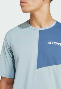 Maglietta sportiva azzurro chiaro con un pannello blu scuro nella parte superiore sinistra, maniche corte e scollo rotondo. Logo "TERREX" bianco.