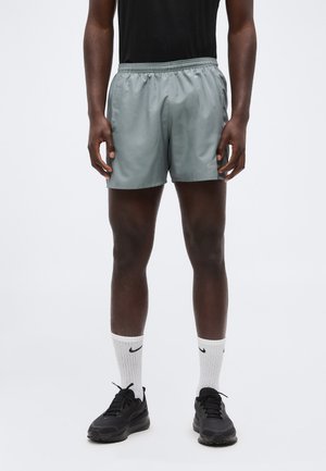 Mann trägt graue Sportshorts, schwarzes Shirt, weiße Nike-Socken und schwarze Turnschuhe, steht vor einem einfarbigen Hintergrund.