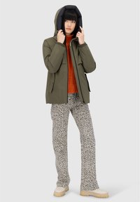 Persona che indossa una giacca con cappuccio verde sopra un maglione arancione, pantaloni con stampa leopardata e stivali beige, mentre aggiusta il cappuccio della giacca su uno sfondo grigio.