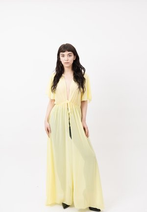 Joven mujer con cabello largo y oscuro vistiendo un vestido fluido de color amarillo claro, de pie frente a un fondo blanco liso.