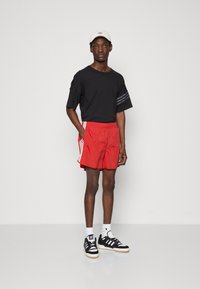 Tricou negru cu mânecă scurtă, cu trei dungi pe mânecă, pantaloni scurți atletici roșii cu dungi albe pe lateral, adidași negri și albi și o șapcă albă.