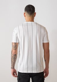 Camiseta blanca de manga corta con rayas verticales negras, cuello redondo y corte ajustado. La tela parece suave con un diseño informal.