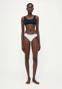 Femme debout pieds nus sur un fond uni, portant un débardeur noir court et des culottes blanches avec une marque "Emporio Armani" visible.