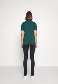 Camisa polo verde de manga corta, con tejido texturizado y cuello plano. Combinada con unos vaqueros skinny negros y unas zapatillas blancas de plataforma, vista desde la parte trasera.