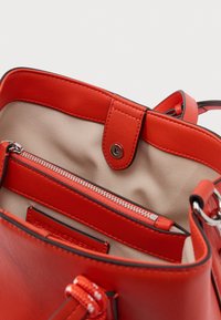 Punainen nahkainen käsilaukku, jossa on beige kangasvuori. Siinä on vetoketjullinen osio ja painonappisuljenta, sekä yksityiskohtaiset ompeleet ja koristeet.