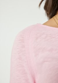 Detail ramene osoby oblečené v světle růžovém pleteném topu a tenkém zlatém řetízku na jednoduchém bílém pozadí.