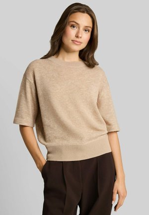 Femme aux longs cheveux bruns portant un pull beige à manches courtes et un pantalon marron foncé, debout avec une main dans la poche sur un fond uni.