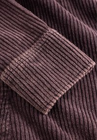 Gros plan sur un tissu en velours côtelé violet texturé montrant des côtes verticales et une section de poignet pliée.
