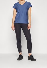 Mujer vistiendo una blusa azul de manga corta y escote en V, leggins negros y zapatillas deportivas negras, de pie contra un fondo claro y liso.