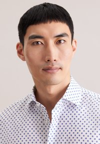 Camicia bianca con pois blu, colletto abbottonato e maniche corte. Tessuto liscio e vestibilità su misura. Primo piano della parte superiore del corpo di un modello maschile.