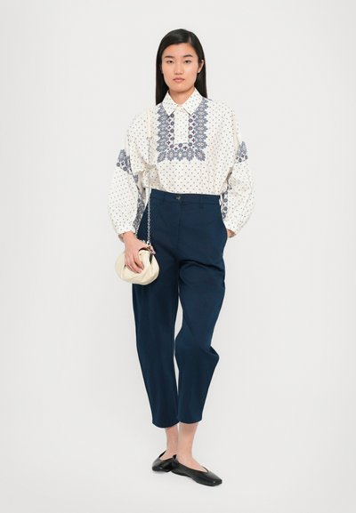 Blusa branca com bordado azul e bolinhas, combinada com calças escuras de cintura alta e sapatos pretos planos, segurando uma bolsa bege.