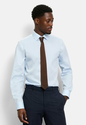 Hellblaues Langarmhemd mit klassischem Kragen, kombiniert mit einer strukturierten braunen Krawatte. Dunkel kariierte Hose vervollständigt das Outfit.
