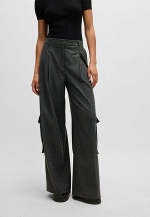 Femme portant un pantalon large gris foncé avec des plis et de grandes poches latérales, assorti à un top noir moulant à manches courtes.