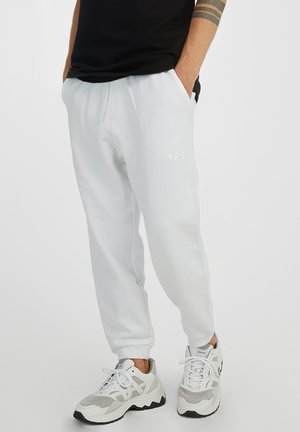 Witte joggingbroek met zijzakken en een trekkoord, voorzien van een subtiele "YPS"-logo in het wit op de linkerdij. Draagt samen met witte en grijze sneakers.