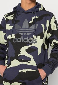 Bluza z kapturem w kamuflażowym wzorze z granatowym, czarnym i jasnooliwkowym kolorem. Posiada logo na klatce piersiowej oraz kieszeń z przodu. Materia ł mieszany bawełniany.