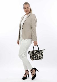 Tasche mit Leopardenmuster und schwarzen Griffen, mit Reißverschlussfächern und strukturiertem Design. Das Model trägt ein beiges Blazer, ein weißes Oberteil und weiße Hosen.