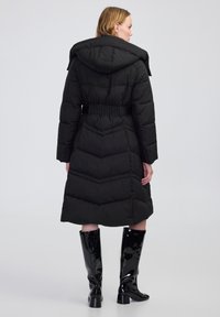 Cappotto imbottito nero con cappuccio, caratterizzato da cuciture a zig-zag orizzontali, vita elastica e indossato con stivali neri lucidi al ginocchio.