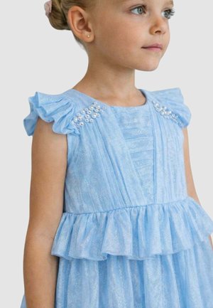 Ragazza giovane che indossa un vestito azzurro chiaro con balze e dettagli di perle sulle spalle, che guarda verso destra su uno sfondo semplice.