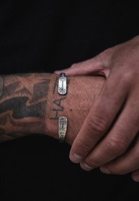 Duos sidabrinių apyrankių ant nuskaitytos, tatuiruotos rankos. Viena turi palmės medžio dizainą; kita turi linijinį raštą.