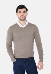 Giovane uomo con capelli scuri e barba che indossa un maglione beige a scollo a V sopra una camicia bianca con colletto e jeans blu scuro, in piedi contro uno sfondo bianco.