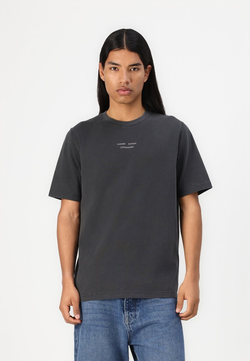 T-shirt in cotone grigio scuro con maniche corte, scollatura rotonda e dettagli testuali sul davanti. Tessuto morbido e vestibilità rilassata. Indossato con jeans in denim blu.
