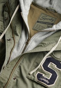 Olive zielona kurtka z szarym wnętrzem z bluzy, z złotym zamkiem i teksturowaną granatową literą "S" naszytą na kieszeni z przodu.