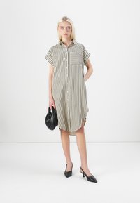 Rochie cămașă cu mânecă scurtă, dungată, în nuanțe de bej și negru, având un decolteu cu nasturi, un singur buzunar pe piept și fante laterale. Poartă accesorii negre.