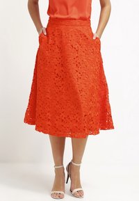 Orange spetskjol med blommig utskärning, utsvängd silhuett och fickor. Har en midja och textil med struktur. Stylad med vita klacksandaler.