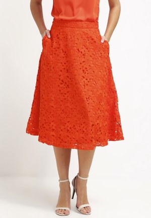 Gonna arancione in pizzo con ritagli floreali, silhouette svasata e tasche. Caratterizzata da una vita alta e un tessuto testurizzato. Abbinata a sandali bianchi con tacco.