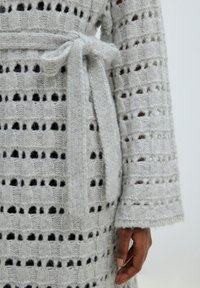 Grijze gebreide cardigan met een opengewerkte patroon, voorzien van een strikceintuur in de taille en lange mouwen met een gestructureerde afwerking en losse manchetten.
