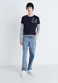 Mørkeblå kortærmet skjorte med stribede lange ærmer, nummer 24 foran; lyseblå jeans; hvide sneakers med grå accenter.