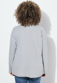 Grau langärmliges Oberteil aus weichem Stoff, mit einem leicht abgerundeten Saum und kontrastierenden Nähten. Getragen mit Jeans.