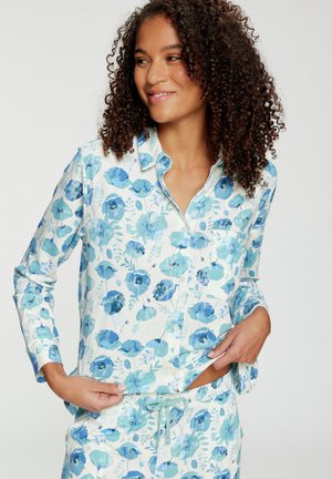 Frau mit lockigem Haar, die einen weißen Pyjama mit blauen Blumenmustern trägt, richtet den Saum des Hemdes und lächelt leicht seitlich.