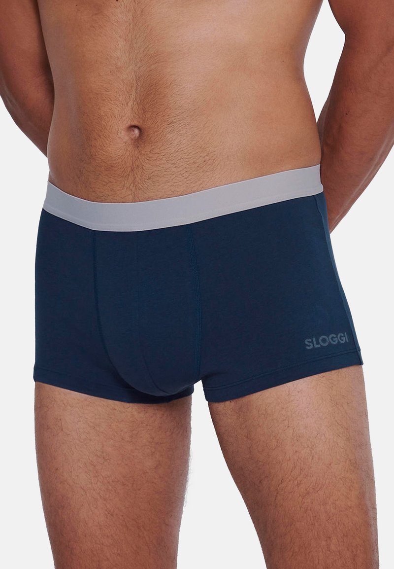 Torso masculin portant un boxer bleu marine avec une ceinture grise claire, arborant le nom de la marque "Sloggi" sur la cuisse gauche.