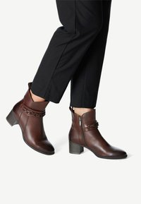 Hnědé kožené kotníkové boty s nízkým fasetovým podpatkem, které mají dekorativní pletené detaily a boční zip pro snadné obouvání.