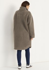 Langer, übergroßer brauner Fleece-Mantel mit hohem Kragen, weicher Textur und lässiger Passform. Kombiniert mit dunklen Jeans und weißen Sneakers.