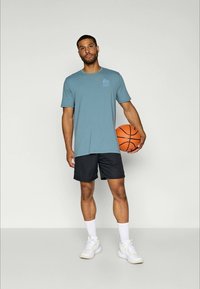 Vīrietis zilā T-kreklā, melnās šortos, baltās zeķēs un sporta apavos stāvot, turot oranžu basketbola bumbu pret vienkāršu fona.